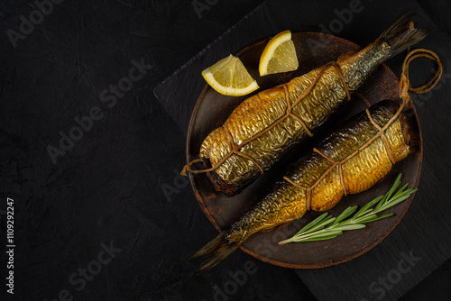 two hot smoked herrings with ropes and spices lie side by side on a clay plate on a stone cutting board and a black background. view from above with copy space