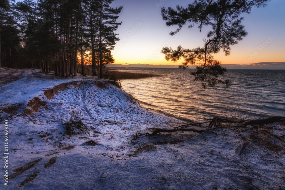 Evening sunset over a large lake with a wave and a high snow-covered coast with a cliff, basin and pine trees. Beautiful winter landscape. side view from the shore