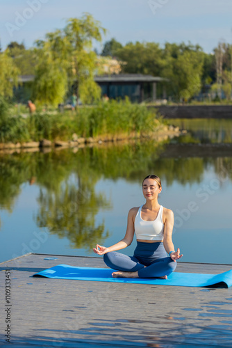 Foto Young woman sitting on mat in lotus position, relaxing near pond in park on summer day