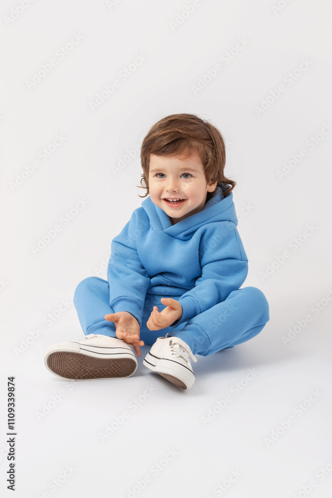 Naklejka premium A Caucasian curly-haired toddler boy of 3 years old is sitting on the floor, smiling and looking attentively with interest in a blue tracksuit on a white background. Vertical photo. Copy Space