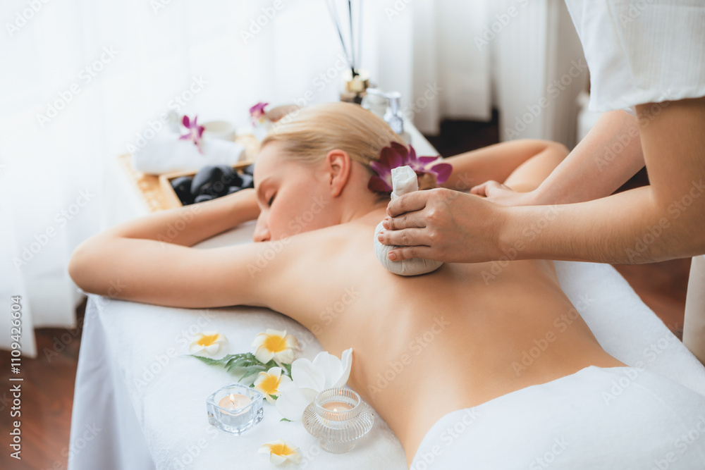 Naklejka premium Hot herbal ball spa massage body treatment, masseur gently compresses herb bag on woman body. Tranquil and serenity of aromatherapy recreation in day lighting ambient at spa salon. Quiescent