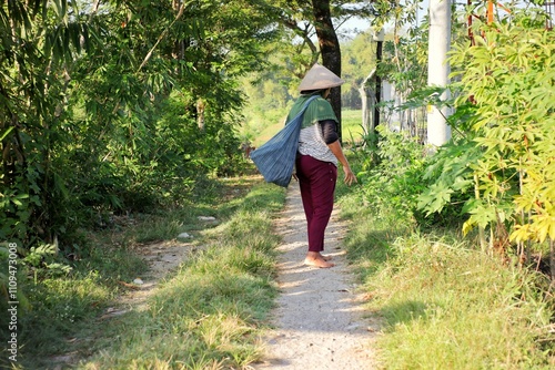 A woman wearing a caping or cone-shaped hat made of woven bamboo while carrying a basket made of cloth on her back, heading to the fields. photo