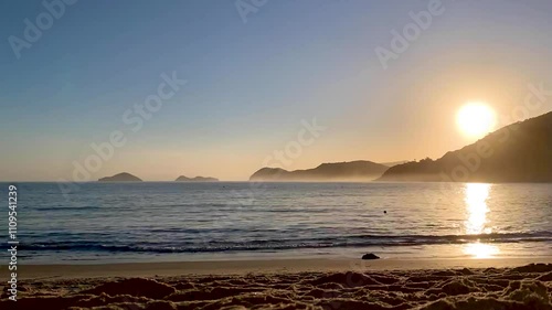 Time-lapse do sol se pondo atrás da montanha e seus raios refletindo no mar. Várias pessoas, bicicletas, cachorros passando, aparecendo apenas suas silhuetas. Ilha das Couve e As Ilhas ao fundo. photo