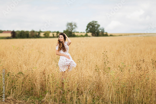 A beautiful brunette woman in a white dress runs along a field of golden chickpeas ear. Stylish girl in the field in windy weather