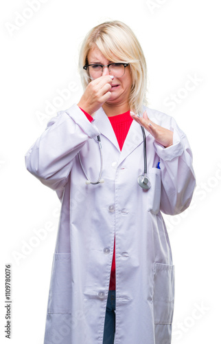 Young beautiful blonde doctor woman wearing medical uniform over isolated background smelling something stinky and disgusting, intolerable smell, holding breath with fingers on nose. Bad smells. photo