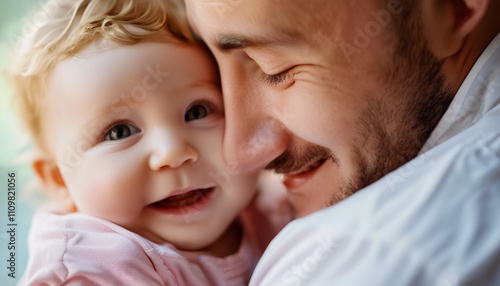 A father holding his baby, both smiling with joy. The baby has blonde hair and is wearing a light