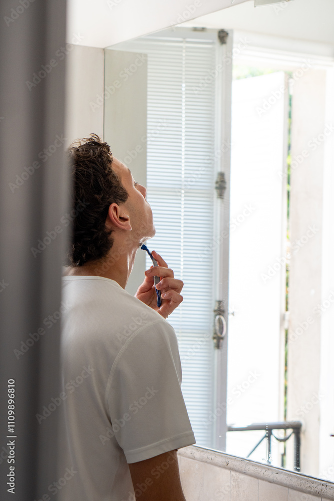 A young man with dark features shaving his beard using a disposable razor, captured in a side view inside a bathroom.