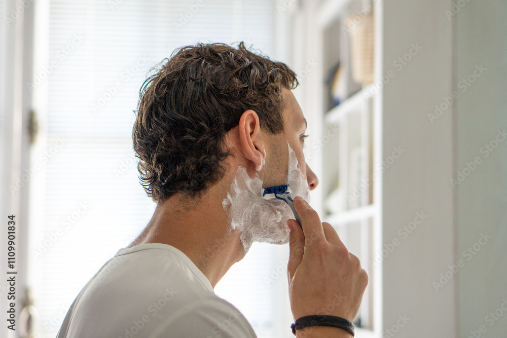 A young man deeply focused while shaving his beard, standing in front of a mirror. A depiction of precision and care in personal grooming.
