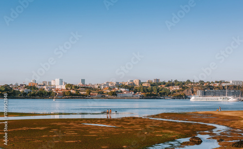 Panoramic view of tourists enjoying the low tide of douro river with the cityscape of Porto in the background, under a clear blue sky, in Vila Nova de Gaia, portugal