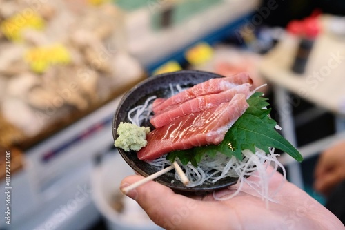 Fatty tuna sashimi slices glazed with soy sauce, and garnished with a perilla leaf and fresh wasabi, held in a woman’s hand at Tsukiji Outer Fish Market in Chuo, Tokyo, Japan 