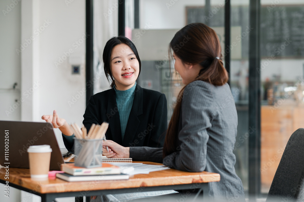 Two women engaged in a business discussion at a modern office table with a laptop and coffee. Two women engaged in a business discussion at a modern office table with a laptop and coffee.