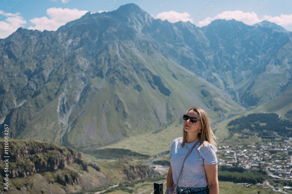 Naklejka premium A tourist girl against the background of the mountains and the village of Stepantsminda in Georgia. The girl is wearing a purple T-shirt, denim shorts, sunglasses and a purple bag. Space, gorgeous vie