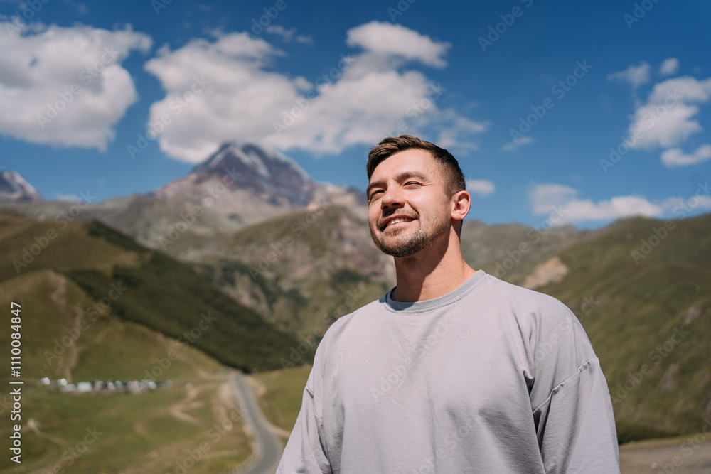 Man standing in the mountains near Kazbek in Georgia, basking in sunlight with a serene expression, with green valleys, rugged peaks, and a clear blue sky in the background Man standing in the mountains near Kazbek in Georgia, basking in sunlight with a serene expression, with green valleys, rugged peaks, and a clear blue sky in the background
