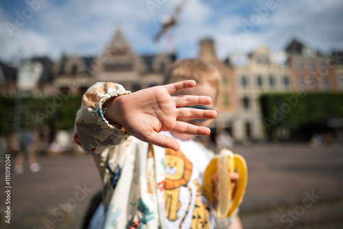 A toddler eating a banana playfully gestures no to photos with their hand, set against a background of defocused Dutch buildings, capturing a candid and lively moment
