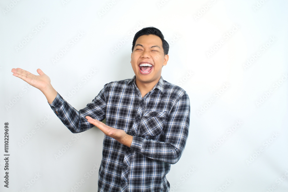 carefree asian man presenting hand with laughing expression showing joy and playful emotion wearing casual plaid shirt isolated on white background