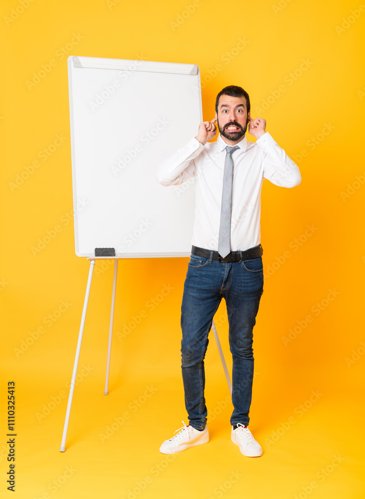 Full-length shot of businessman giving a presentation on white board over isolated yellow background frustrated and covering ears Full-length shot of businessman giving a presentation on white board over isolated yellow background frustrated and covering ears