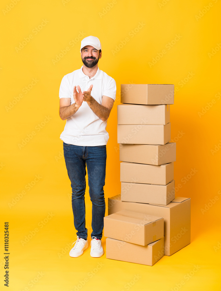 Full-length shot of delivery man among boxes over isolated yellow background applauding after presentation in a conference Full-length shot of delivery man among boxes over isolated yellow background applauding after presentation in a conference