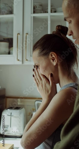 A woman is crying standing in the kitchen after a family quarrel. Her husband hugs her from behind trying to make peace