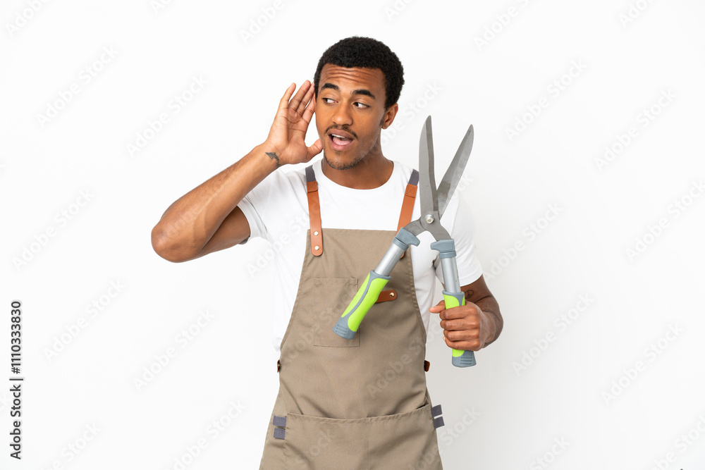 African American gardener man holding pruning shears over isolated white background listening to something by putting hand on the ear African American gardener man holding pruning shears over isolated white background listening to something by putting hand on the ear