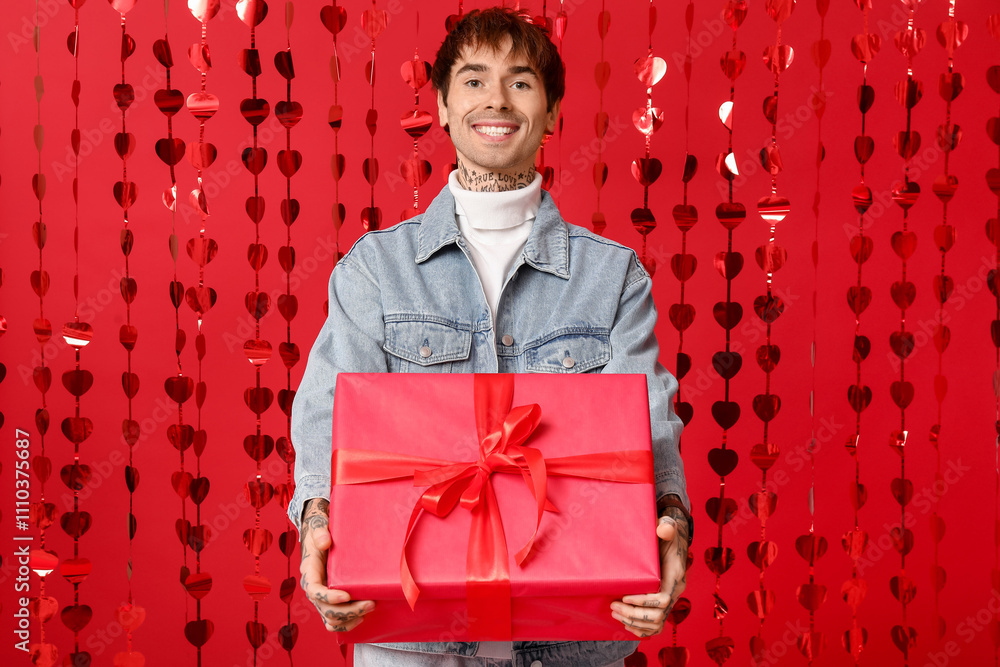Handsome young man with gift box and hearts curtain on red background. Valentine's Day celebration Handsome young man with gift box and hearts curtain on red background. Valentine's Day celebration