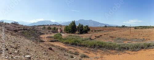 Moroccan countryside scene: red earth, sparse vegetation, a pink house, and distant mountains under a serene sky.