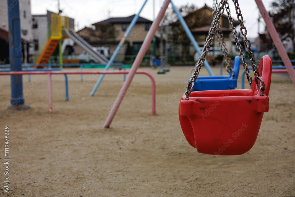 In a typical Japanese inner city neighborhood playground, red and blue infant swings sit silent on an overcast day. The bokeh background features a colorful playground slide and neighborhood homes. In a typical Japanese inner city neighborhood playground, red and blue infant swings sit silent on an overcast day. The bokeh background features a colorful playground slide and neighborhood homes.