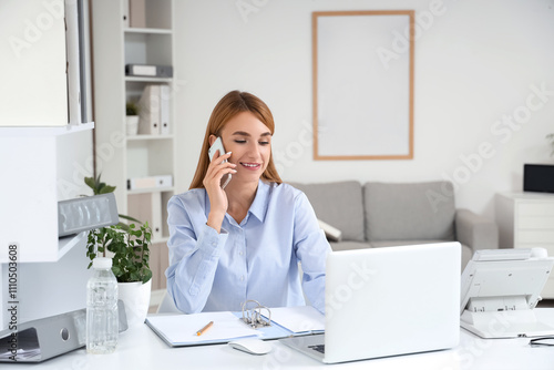 Young businesswoman with laptop talking by mobile phone at desk in office