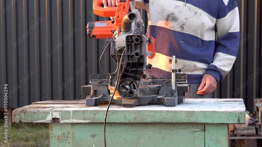 A man works with a construction tool. Table saw for wood. In the background is a house. Outdoors. Construction work. Tool. Close-up. A man works with a construction tool. Table saw for wood. In the background is a house. Outdoors. Construction work. Tool. Close-up.