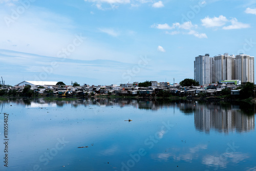 A mix of small, densely packed houses and tall modern buildings reflecting in a body of water under a clear blue sky, highlighting urban contrast and development.