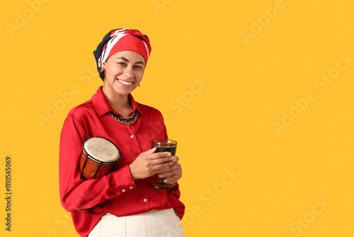 Young African-American woman with drink and tam-tam drum on yellow background. Kwanzaa celebration photo