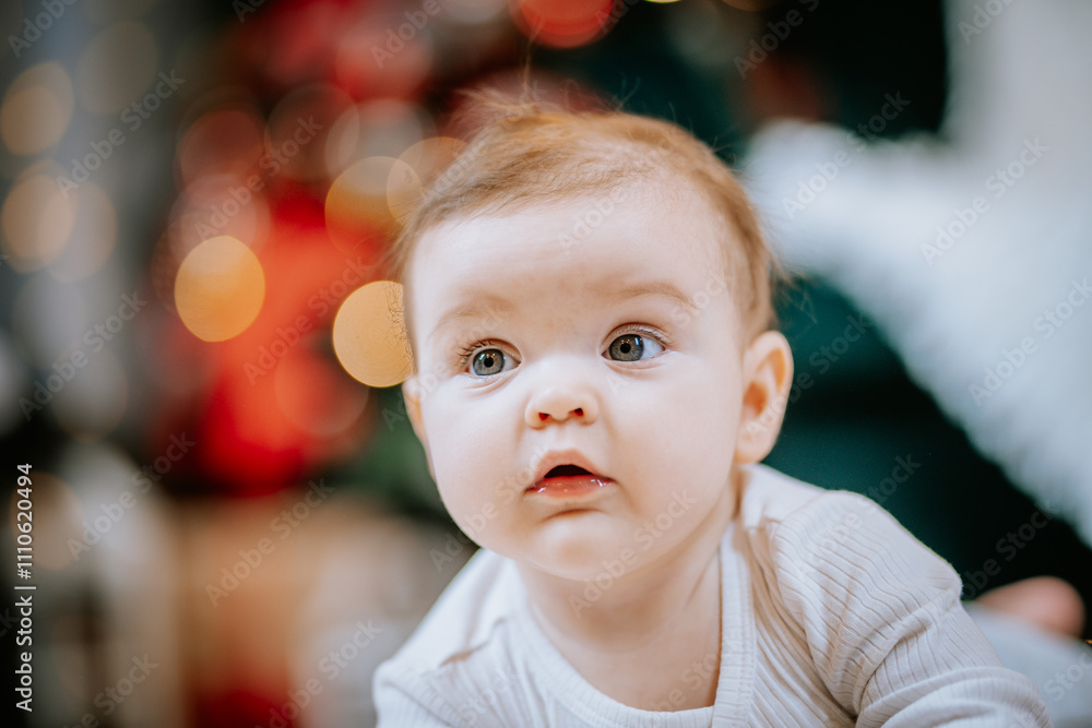 Close-up of a baby with wide eyes and a curious expression, with blurred festive Christmas lights and decor in the background. Close-up of a baby with wide eyes and a curious expression, with blurred festive Christmas lights and decor in the background.