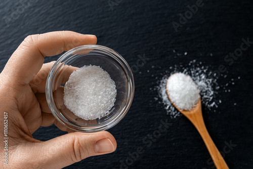 Bowl of monosodium glutamate in woman fingers over black background. Crystals of flavor enhancer MSG in a glass container. Food additive E621 concept. photo