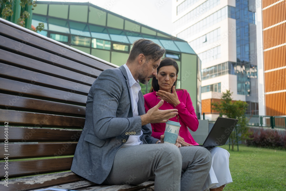 A business man and woman sitting on a bench in a park, in front of a complex of modern glass office buildings. They are having a discussion, exchanging ideas while using a laptop. A business man and woman sitting on a bench in a park, in front of a complex of modern glass office buildings. They are having a discussion, exchanging ideas while using a laptop.