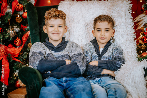 Two brothers sit together on a cozy chair, smiling in a festive Christmas setting with decorated trees, lights, and holiday decorations. photo