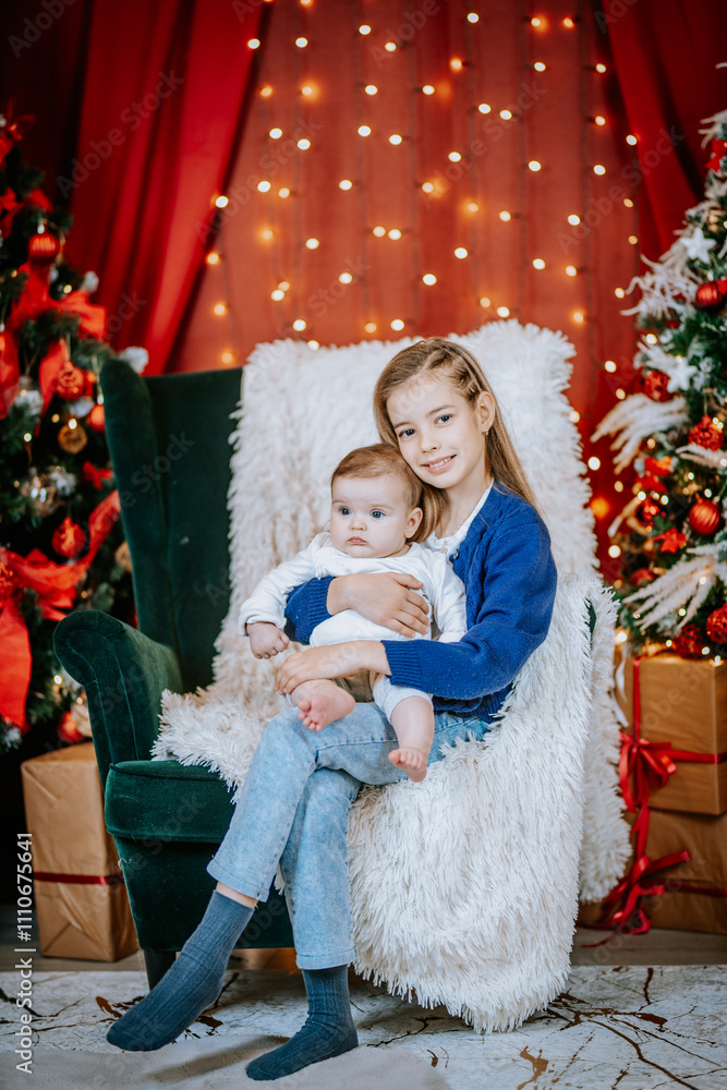 Obraz premium A young girl holds a baby on a cozy chair surrounded by festive Christmas lights, decorated trees, and wrapped gifts in a warm holiday setting.
