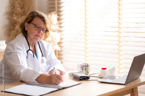 Senior woman doctor in white coat with stethoscope working with writing notes of patient while laptop on desk in modern medical office, female doctor diagnosis with concentration in clinic.