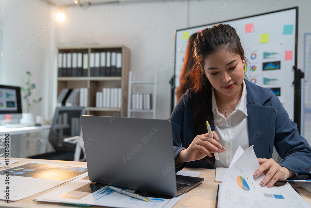 Asian businesswoman reviewing financial documents and using laptop, working with graphs and data in modern office, focused on analysis and planning