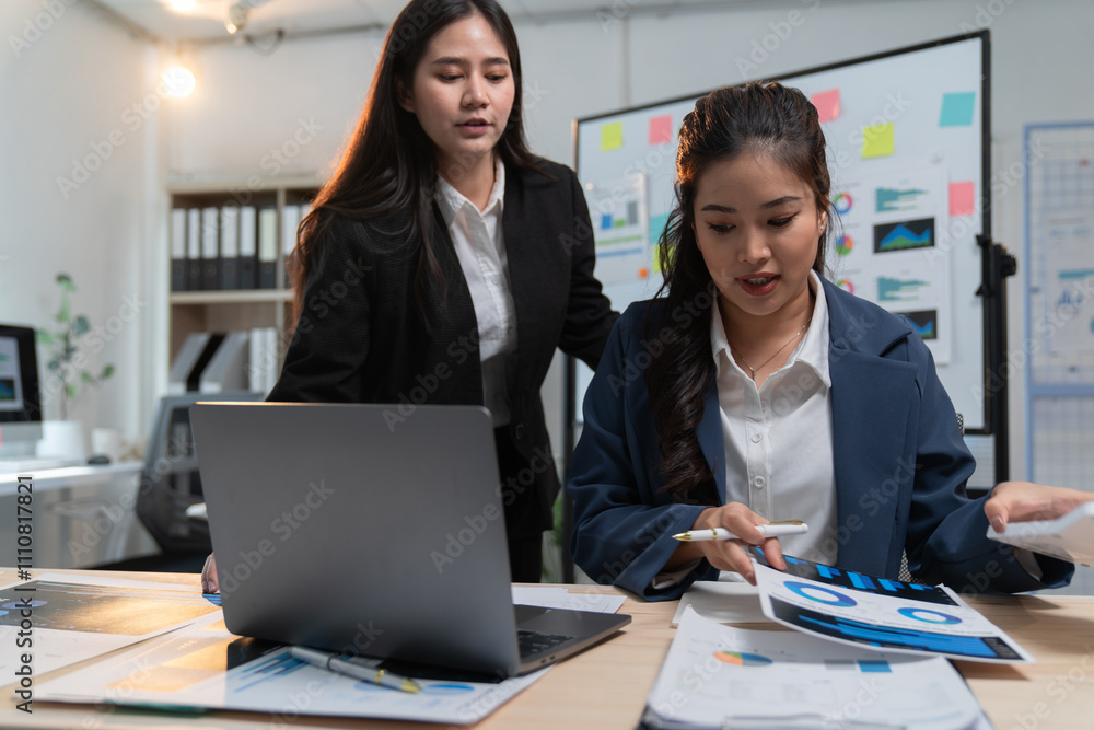 Two asian businesswomen are collaborating on analyzing financial reports, using a laptop and printed charts in a well-lit office environment Two asian businesswomen are collaborating on analyzing financial reports, using a laptop and printed charts in a well-lit office environment
