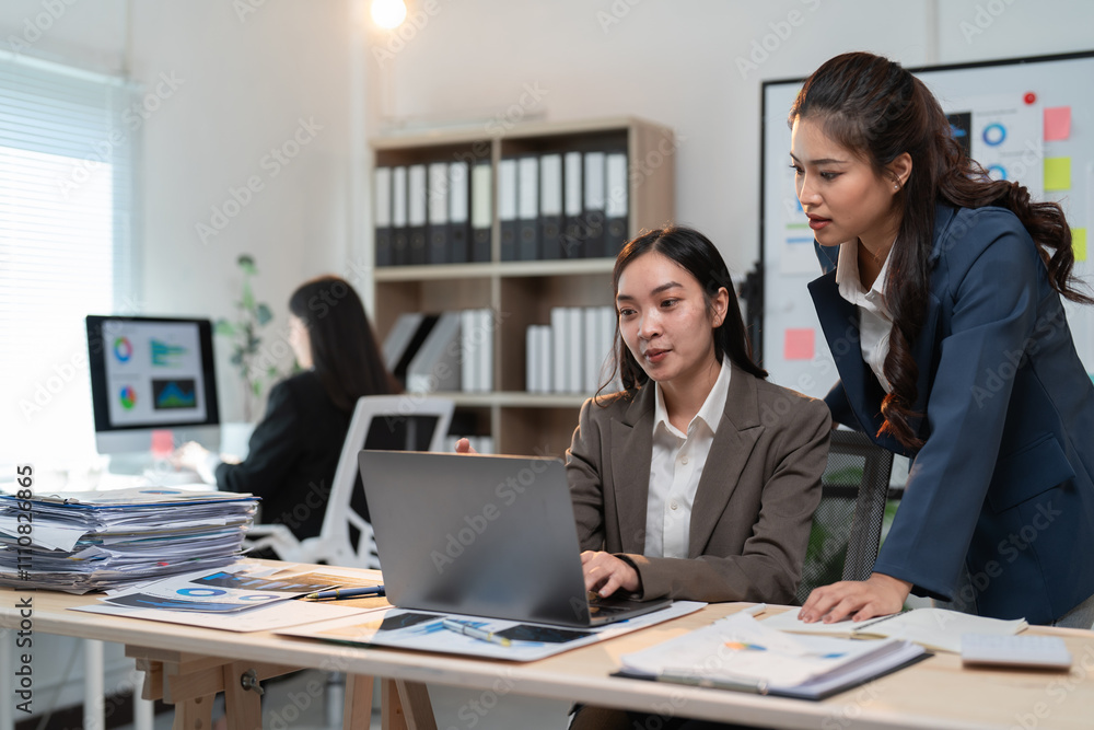 Two asian businesswomen are working together in a modern office, focusing on a laptop and analyzing data while another colleague works in the background Two asian businesswomen are working together in a modern office, focusing on a laptop and analyzing data while another colleague works in the background