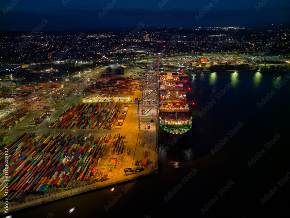 Illuminated Southampton UK container terminal with large container ship at night. High altitude aerial view above bright commercial docks towards the city. Bow of the vessel.