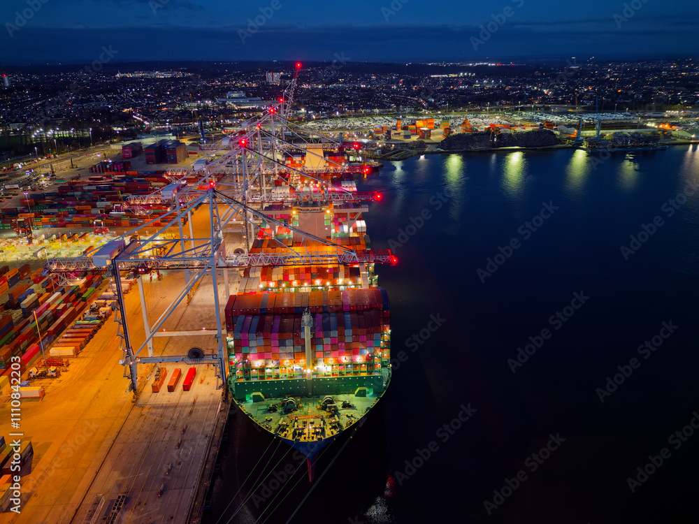 Above bow of large container ship at Southampton Docks UK aerial view. Illuminated container terminal with cranes and lights reflection on calm sea. Beautiful cargo and shipping industrial background.