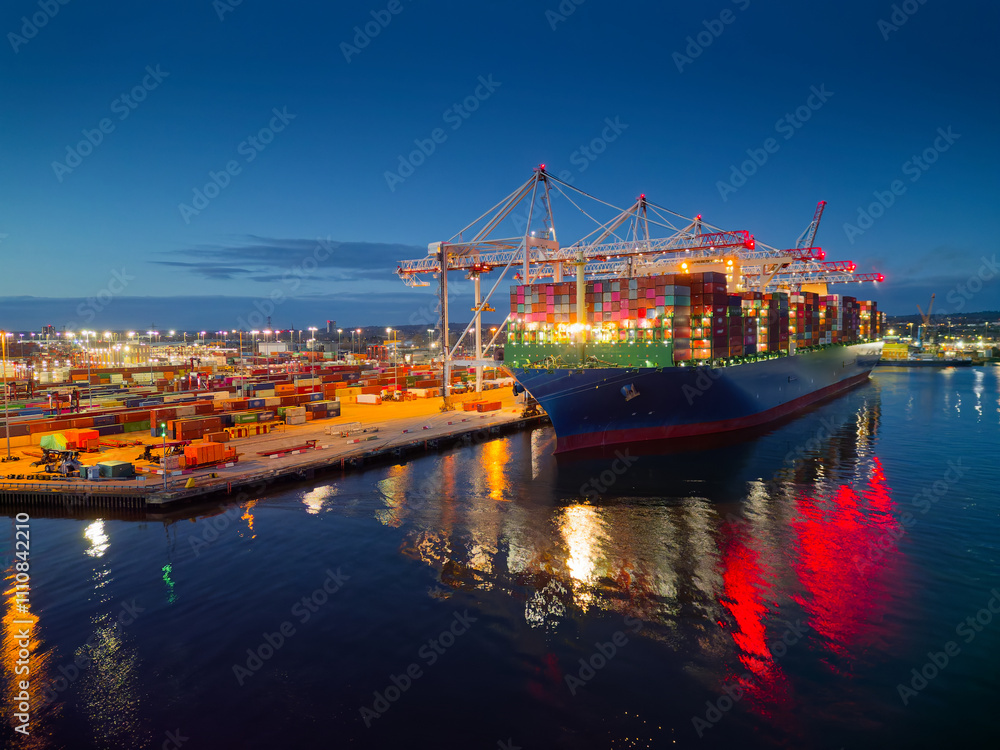 Illuminated Southampton UK container terminal with large container ship at dusk. Beautiful light reflection on calm sea and dark blue sky. Place of work in shipping industry.
