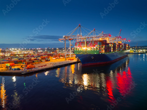 Obraz na płótnie Illuminated Southampton UK container terminal with large container ship at dusk. Beautiful light reflection on calm sea and dark blue sky. Place of work in shipping industry. w salonie