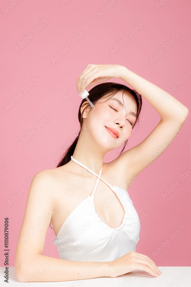 Young Asian woman gathered in ponytail with natural makeup have plump lips and clean fresh skin wearing white camisole applying moisturizing serum on face on isolated pink background. Young Asian woman gathered in ponytail with natural makeup have plump lips and clean fresh skin wearing white camisole applying moisturizing serum on face on isolated pink background.