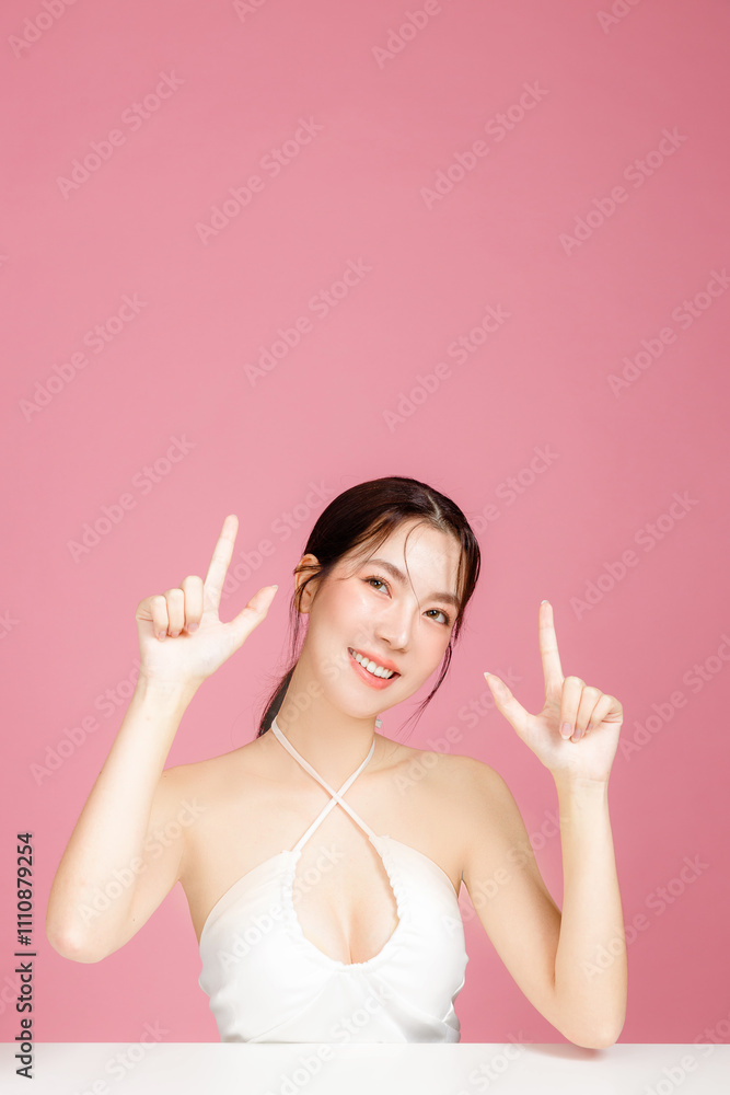 Young Asian woman gathered in ponytail with natural makeup on face have plump lips and clean fresh skin wearing white camisole pointing with finger to copy space on isolated pink background. Young Asian woman gathered in ponytail with natural makeup on face have plump lips and clean fresh skin wearing white camisole pointing with finger to copy space on isolated pink background.