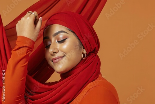 A young woman wearing a vibrant red hijab and matching outfit poses in front of a warm orange background. She gracefully interacts with flowing fabric, showcasing confidence and beauty in her expressi photo