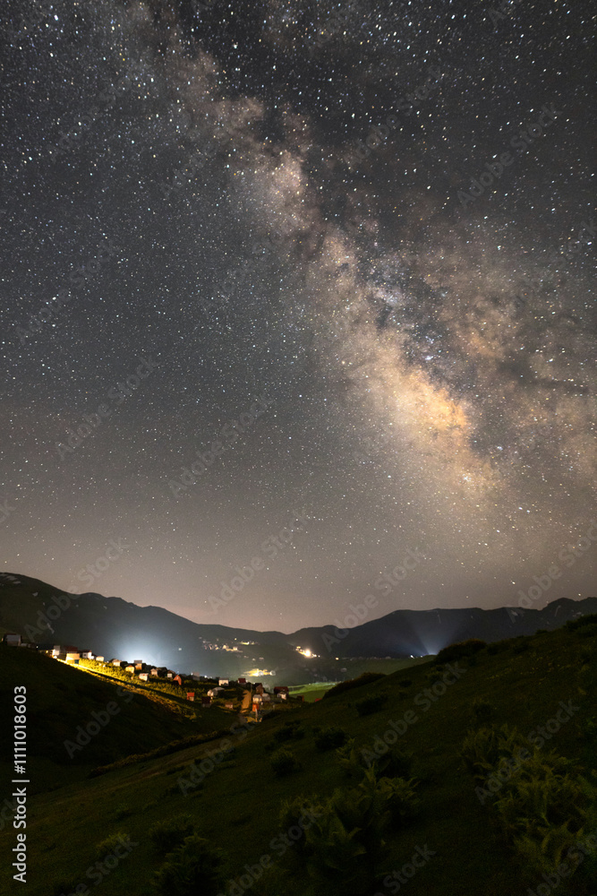 A night sky with the Milky Way galaxy visible, a small village illuminated below, and hills in the landscape A night sky with the Milky Way galaxy visible, a small village illuminated below, and hills in the landscape