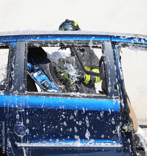 firefighter stands behind a destroyed blue car while spraying a fire extinguishing foam to put out the smoldering remains photo