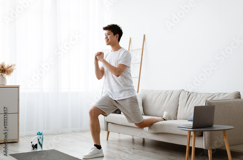 Online Training. Smiling Sporty Asian Guy Doing Single-Leg Split Squats Near Couch Exercising Using Laptop Computer During Workout At Home. Domestic Male Fitness And Sporty Lifestyle. Side View photo