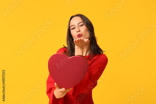 Beautiful young woman blowing kiss with gift box in shape of heart on yellow background. Valentine's Day celebration